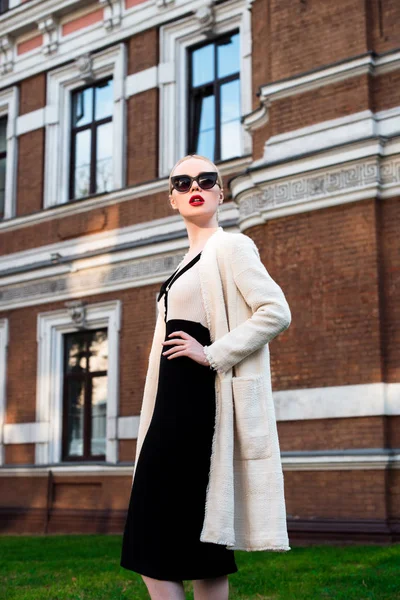 Happy Fashion blonde european elegant Woman with red lips and white skin Standing at the Old Red Brick building on green grass. Glamour Girl Outdoors. Beautiful Female in formal black and white dress