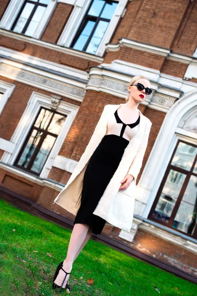 Happy Fashion blonde european elegant Woman with red lips and white skin Standing at the Old Red Brick building on green grass. Glamour Girl Outdoors. Beautiful Female in formal black and white dress