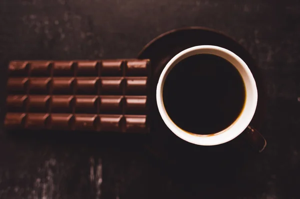 Tasse Kaffee mit Körnern und Schokolade auf Holztisch vor dunklem Hintergrund — Stockfoto