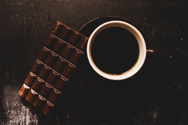 Tasse Kaffee mit Körnern und Schokolade auf Holztisch vor dunklem Hintergrund — Stockfoto