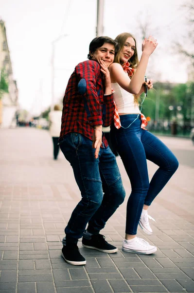 Zwei fröhliche trendige Teenager-Freunde, die durch die Stadt spazieren und tanzen, die Musik mit Kopfhörern hören, miteinander reden und lächeln. Lebensstil, Freundschaft und urbane Lebenskonzepte. — Stockfoto