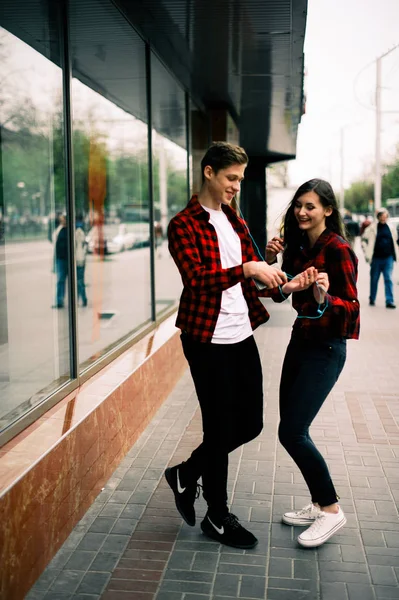 Deux amis adolescents joyeux et branchés marchent et dansent en ville, écoutent la musique avec des écouteurs, se parlent et sourient. Style de vie, amitié et concepts de vie urbaine . — Photo