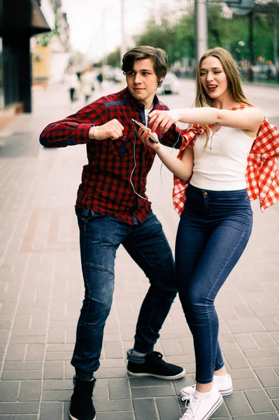 Due allegri amici adolescenti alla moda che passeggiano e ballano in città, ascoltano la musica con le cuffie, si parlano e sorridono. Stile di vita, amicizia e concetti di vita urbana . — Foto Stock