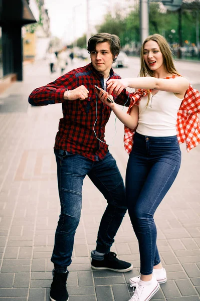 Dois felizes amigos adolescentes da moda andando e dançando na cidade, ouvindo a música com fones de ouvido, conversando uns com os outros e sorrindo. Estilo de vida, amizade e conceitos de vida urbana . — Fotografia de Stock