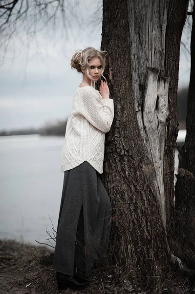 Красива молода блондинка з блакитними очима в білому пуловері довга спідниця на весняному тлі, лісі, озері, деревах. Холодний настрій. Мода, емоційний портрет — стокове фото