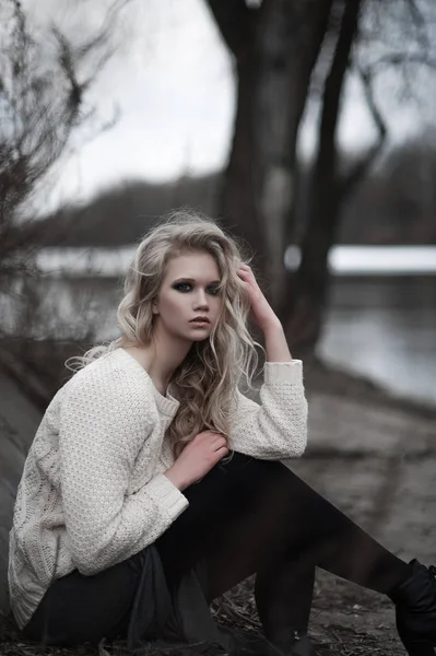 Mooie jonge blonde vrouw met blauwe ogen in de witte trui lange rok op voorjaar achtergrond, bomen, bos, lake. Koude sfeer. Mode, emotionele portret — Stockfoto