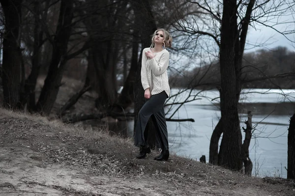 Vacker ung blond kvinna med blå ögon i vit tröja lång kjol på våren bakgrund, skogen, sjön, träd. Kall humör. Mode, känslomässiga porträtt Stockfoto