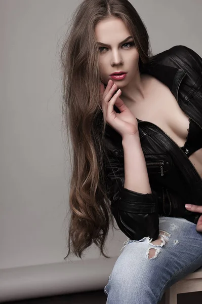 Giovane ragazza con lunghi bei capelli e occhi fumosi indossa giacca di pelle nera e jeans seduti sulla sedia. Studio girato . — Foto Stock