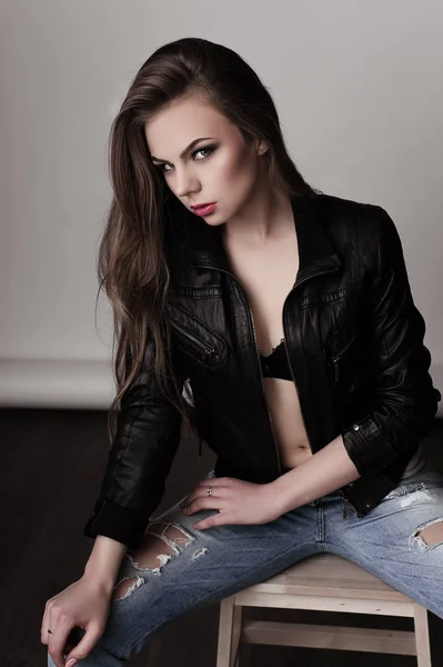 Chica joven con el pelo largo y hermoso y ojos ahumados usando chaqueta de cuero negro y vaqueros sentados en la silla. Captura de estudio . —  Fotos de Stock