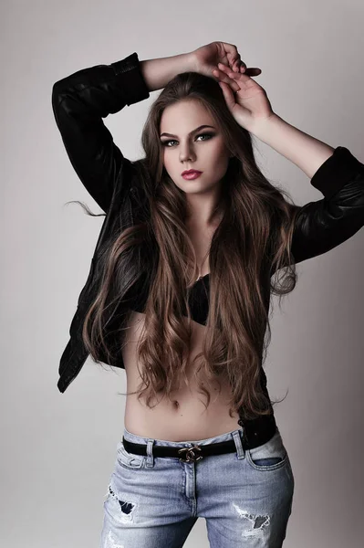 Jeune fille aux longs beaux cheveux et aux yeux fumés portant une veste et un jean en cuir noir. Plan studio . — Photo