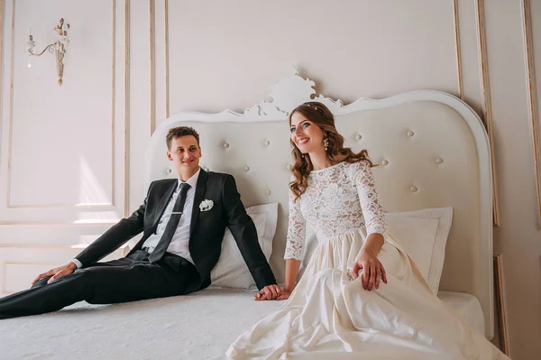 Cute wedding couple in the interior of a classic white studio posing on a bed decorated. hey kiss and hug each other, holding hands looking at each other — Stock Photo, Image