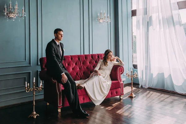 Casal de casamento bonito no interior de um estúdio clássico posando no sofá. Ei beijar e abraçar uns aos outros, de mãos dadas olhando um para o outro — Fotografia de Stock