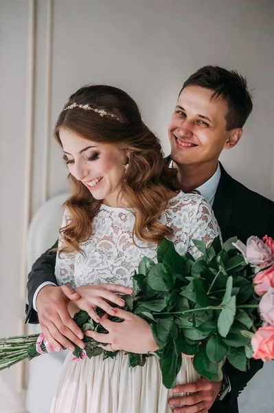 Casal de casamento bonito no interior de um estúdio branco clássico decorado. Ei beijar e abraçar uns aos outros, de mãos dadas olhando um para o outro — Fotografia de Stock