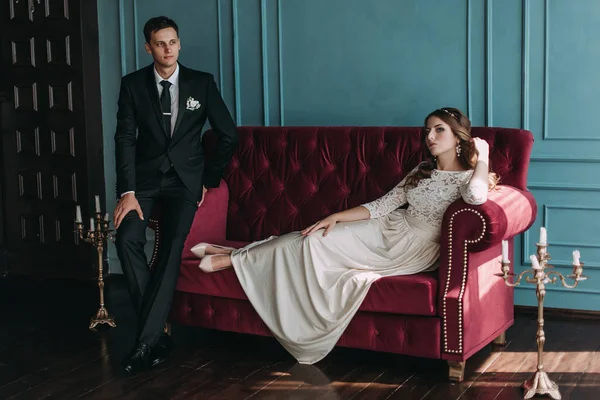 Casal de casamento bonito no interior de um estúdio clássico posando no sofá. Ei beijar e abraçar uns aos outros, de mãos dadas olhando um para o outro — Fotografia de Stock