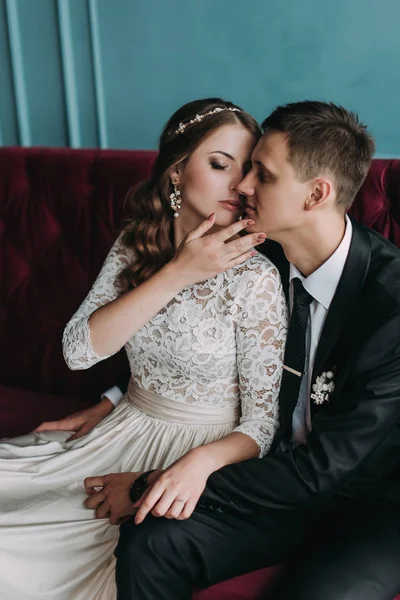 Casal de casamento bonito no interior de um estúdio clássico posando no sofá. Ei beijar e abraçar uns aos outros, de mãos dadas olhando um para o outro — Fotografia de Stock