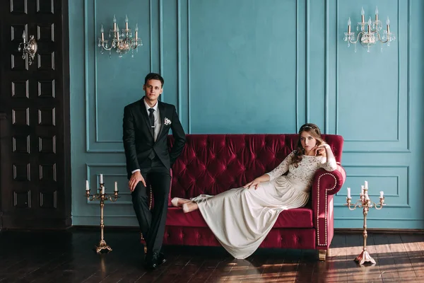 Casal de casamento bonito no interior de um estúdio clássico posando no sofá. Ei beijar e abraçar uns aos outros, de mãos dadas olhando um para o outro — Fotografia de Stock