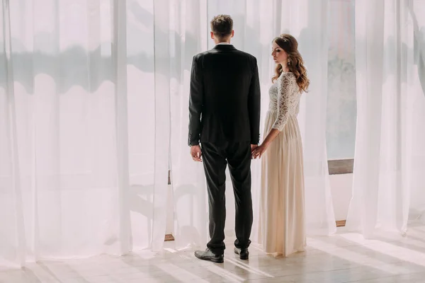 Casal de casamento bonito no interior de um estúdio branco clássico posando em uma cama decorada. Ei beijar e abraçar uns aos outros, de mãos dadas olhando um para o outro — Fotografia de Stock