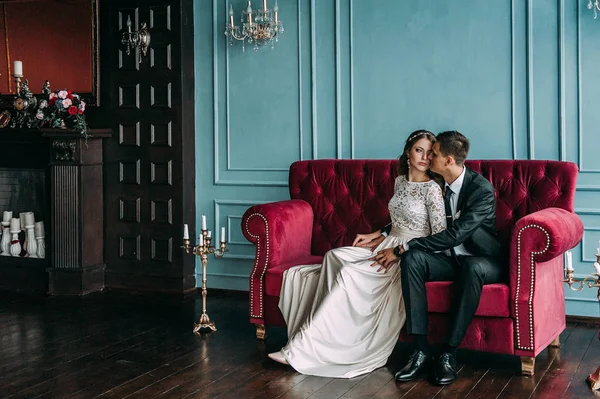Casal de casamento bonito no interior de um estúdio clássico posando no sofá. Ei beijar e abraçar uns aos outros, de mãos dadas olhando um para o outro — Fotografia de Stock