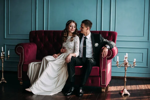 Casal de casamento bonito no interior de um estúdio clássico posando no sofá. Ei beijar e abraçar uns aos outros, de mãos dadas olhando um para o outro — Fotografia de Stock