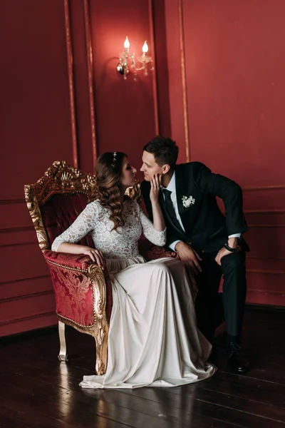 Casal de casamento bonito no interior de um estúdio clássico com fundo vermelho. Ei beijar e abraçar uns aos outros, de mãos dadas olhando um para o outro — Fotografia de Stock