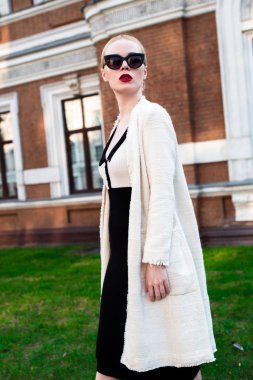 Mutlu moda sarışın Avrupa zarif kadın kırmızı dudakları ve beyaz ayakta yeşil çimenlerin üzerinde bina eski kırmızı tuğla, Cilt. Seksi kız açık havada. Resmi siyah beyaz elbiseli güzel kadın 