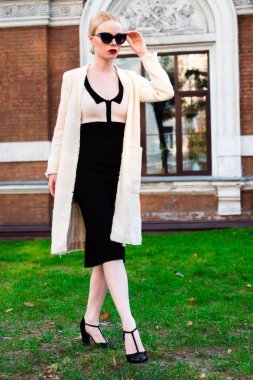 Mutlu moda sarışın Avrupa zarif kadın kırmızı dudakları ve beyaz ayakta yeşil çimenlerin üzerinde bina eski kırmızı tuğla, Cilt. Seksi kız açık havada. Resmi siyah beyaz elbiseli güzel kadın 