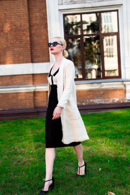 Mutlu moda sarışın Avrupa zarif kadın kırmızı dudakları ve beyaz ayakta yeşil çimenlerin üzerinde bina eski kırmızı tuğla, Cilt. Seksi kız açık havada. Resmi siyah beyaz elbiseli güzel kadın 