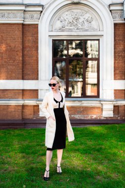 Mutlu moda sarışın Avrupa zarif kadın kırmızı dudakları ve beyaz ayakta yeşil çimenlerin üzerinde bina eski kırmızı tuğla, Cilt. Seksi kız açık havada. Resmi siyah beyaz elbiseli güzel kadın 