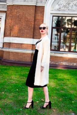 Mutlu moda sarışın Avrupa zarif kadın kırmızı dudakları ve beyaz ayakta yeşil çimenlerin üzerinde bina eski kırmızı tuğla, Cilt. Seksi kız açık havada. Resmi siyah beyaz elbiseli güzel kadın 