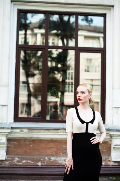 Happy Fashion blonde european elegant Woman with red lips and white skin Standing at the Old Red Brick building on green grass. Glamour Girl Outdoors. Beautiful Female in formal black and white dress.