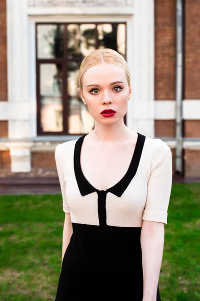 Happy Fashion blonde european elegant Woman with red lips and white skin Standing at the Old Red Brick building on green grass. Glamour Girl Outdoors. Beautiful Female in formal black and white dress.