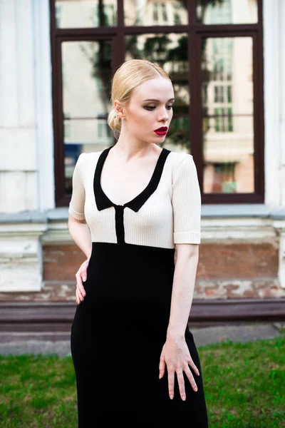 Happy Fashion blonde european elegant Woman with red lips and white skin Standing at the Old Red Brick building on green grass. Glamour Girl Outdoors. Beautiful Female in formal black and white dress.