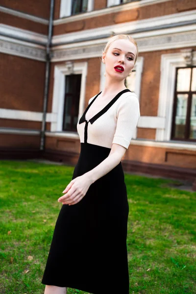 Happy Fashion blonde european elegant Woman with red lips and white skin Standing at the Old Red Brick building on green grass. Glamour Girl Outdoors. Beautiful Female in formal black and white dress.