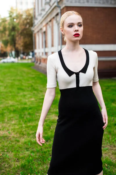 Mutlu moda sarışın Avrupa zarif kadın kırmızı dudakları ve beyaz ayakta yeşil çimenlerin üzerinde bina eski kırmızı tuğla, Cilt. Seksi kız açık havada. Resmi siyah beyaz elbiseli güzel kadın.