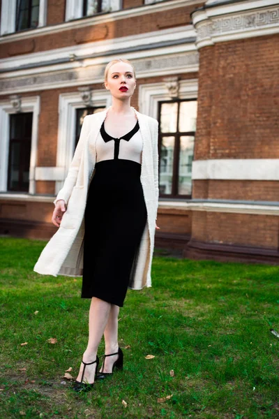 Happy Fashion blonde european elegant Woman with red lips and white skin Standing at the Old Red Brick building on green grass. Glamour Girl Outdoors. Beautiful Female in formal black and white dress.