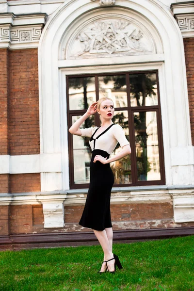 Happy Fashion blonde european elegant Woman with red lips and white skin Standing at the Old Red Brick building on green grass. Glamour Girl Outdoors. Beautiful Female in formal black and white dress.