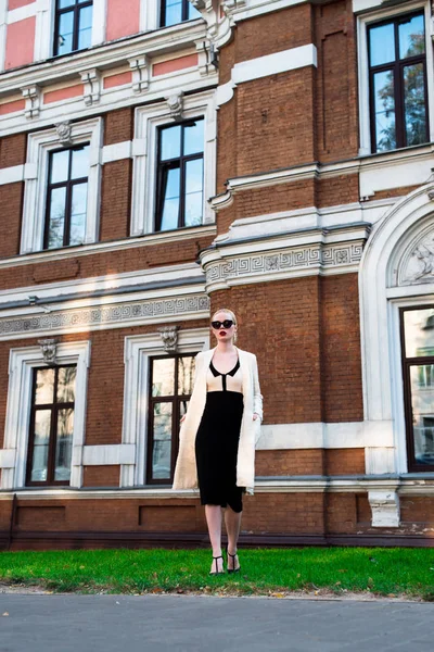 Happy Fashion blonde european elegant Woman with red lips and white skin Standing at the Old Red Brick building on green grass. Glamour Girl Outdoors. Beautiful Female in formal black and white dress