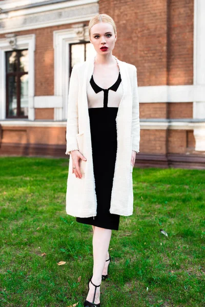 Happy Fashion blonde european elegant Woman with red lips and white skin Standing at the Old Red Brick building on green grass. Glamour Girl Outdoors. Beautiful Female in formal black and white dress.