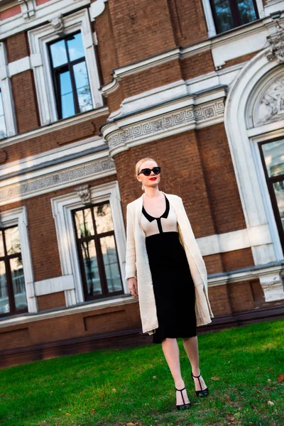 Happy Fashion blonde european elegant Woman with red lips and white skin Standing at the Old Red Brick building on green grass. Glamour Girl Outdoors. Beautiful Female in formal black and white dress