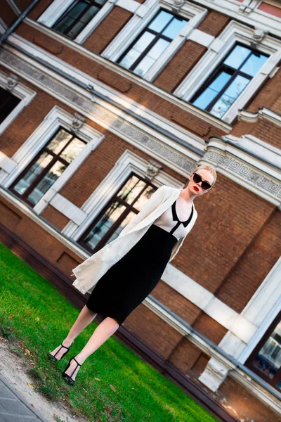 Happy Fashion blonde european elegant Woman with red lips and white skin Standing at the Old Red Brick building on green grass. Glamour Girl Outdoors. Beautiful Female in formal black and white dress