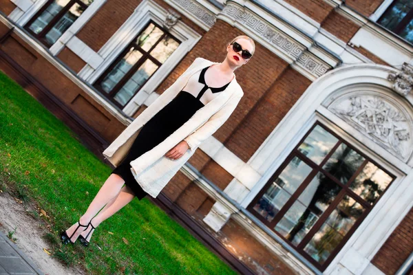 Happy Fashion blonde european elegant Woman with red lips and white skin Standing at the Old Red Brick building on green grass. Glamour Girl Outdoors. Beautiful Female in formal black and white dress