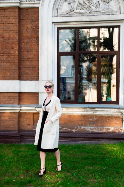 Mutlu moda sarışın Avrupa zarif kadın kırmızı dudakları ve beyaz ayakta yeşil çimenlerin üzerinde bina eski kırmızı tuğla, Cilt. Seksi kız açık havada. Resmi siyah beyaz elbiseli güzel kadın 