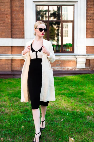 Happy Fashion blonde european elegant Woman with red lips and white skin Standing at the Old Red Brick building on green grass. Glamour Girl Outdoors. Beautiful Female in formal black and white dress