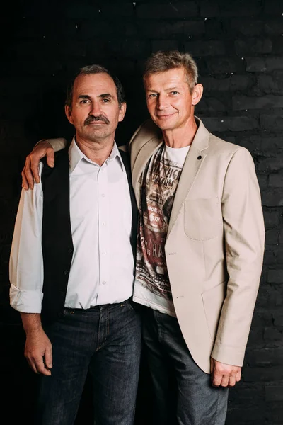 Two handsome middle aged men, one is dressed in a white shirt and a black vest, other wears jacket , old friends. Black background.