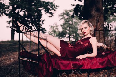 Kırmızı dudaklar ve saç ile güzel ve zarif sarışın kadın giyiyor şarap kırmızı gece sonbahar, retro vintage tarzı ve moda yatakta açık havada poz dalgalar. Düzeltisiz tonlama atış