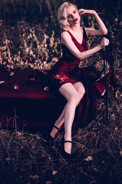 Όμορφο και κομψό ξανθιά γυναίκα με κόκκινα χείλη και τα μαλλιά κύματα φορώντας κρασί κόκκινο Νυχτικό ποζάρει στο κρεβάτι σε εξωτερικούς χώρους το φθινόπωρο, ρετρό vintage στυλ και τη μόδα. Retouched τόνωση βολή — Φωτογραφία Αρχείου