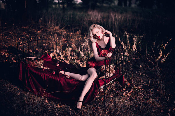 Beautiful and elegant blonde woman with red lips and hair waves wearing wine red nightie posing on the bed outdoors, retro vintage style and fashion