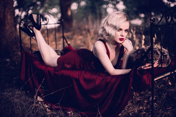 Vacker och elegant blond kvinna med röda läppar och hår vinkar bär vin rött nattlinne poserar på sängen utomhus höst, retro vintage stil och mode. Retuscheras toning skott — Stockfoto