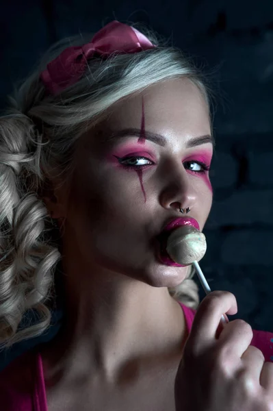 Mooie blonde meisje met twee pigtails, met creatieve pop make-up: roze glanzend lippen, het dragen van roze skelet jurk likken lollipop. voor de Halloween-party. Close-up, geretoucheerde foto. — Stockfoto