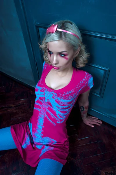 Vacker blond tjej med två flätor, med kreativa docka make-up: rosa glansiga läppar, bär Rosa skelett klänning, poserar nära dörren, sitter på golvet. för Halloween-festen. Poserar som en d — Stockfoto